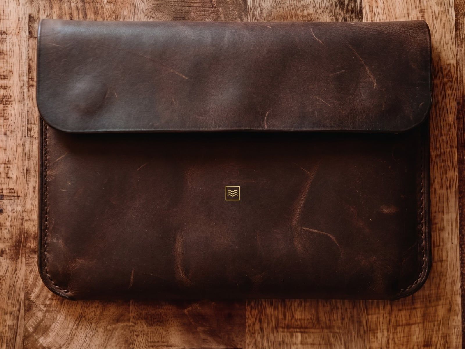 A closed sleeve in dark crazy horse leather with a flap, stitched sides and the Tidemark monogram in gold foil.